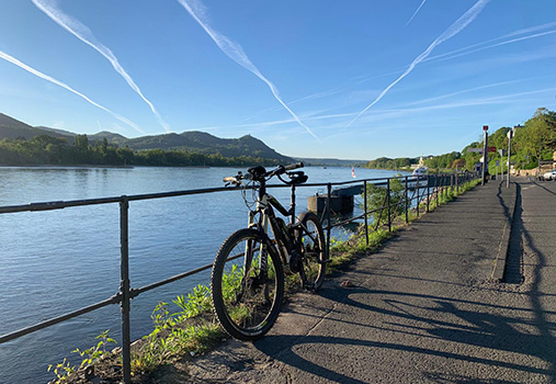 Fahrradtour Fahrrad am Rheinufer auf einem befestigten Uferweg. Im Hintergrund sind der Rhein, bewaldete Hügel und ein klarer Himmel mit Kondensstreifen zu sehen. Die Aufnahme vermittelt den Eindruck einer Fahrradtour entlang des Flusses bei sonnigem Wetter.